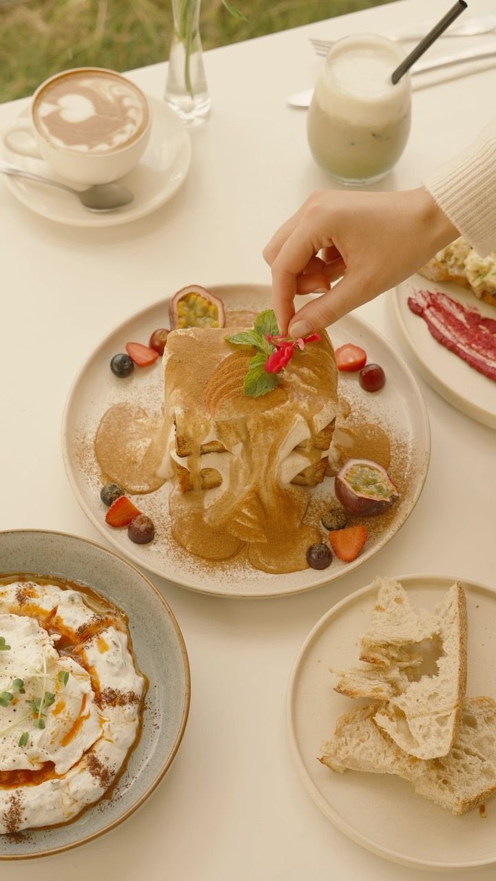 Stacked toast with cream, sauce and fresh fruit, beside yoghurt, bread and coffee